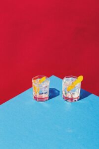 Zwei Gläser mit Eiswürfeln und Orangenschalen auf einem blauen Tisch, vor einem roten Hintergrund. Ideal für Cocktailrezepte und Getränkedekorationen.