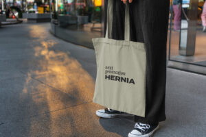 Beige Baumwolltasche mit dem Aufdruck "HERNIA", getragen von einer Person in schwarzer Kleidung, auf einem urbanen Gehweg bei Sonnenuntergang.