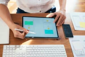 Eine Person arbeitet an einem Tablet, während sie mit einem Stylus Notizen macht. Auf dem Tisch sind eine Tastatur und ein Smartphone zu sehen. Die Bildschirmansicht zeigt zwei grüne Textfelder.