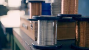 Nahaufnahme einer Spule mit silbernem Draht, umgeben von weiteren Spulen und Werkzeugen in einer Werkstatt. Ideal für Anwendungen in der Elektronik oder Handwerkskunst.