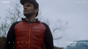 Mann in rot-schwarzer Outdoor-Jacke steht in der Natur und blickt nachdenklich in die Ferne, im Hintergrund ist ein Volkswagen Fahrzeug sichtbar.