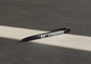 Schwarzer Kugelschreiber mit dem Wort "CONSENS" auf einem grauen Hintergrund, beleuchtet von einem Lichtstrahl.