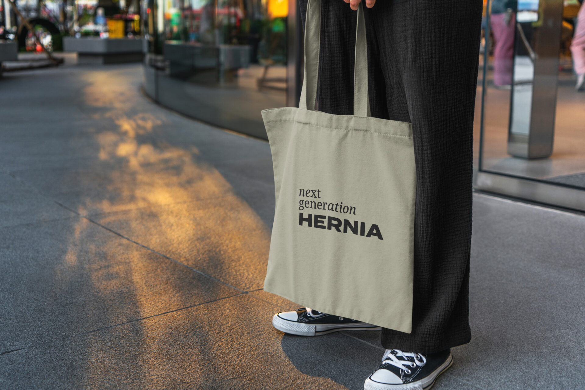 Beige Tragetasche mit dem Schriftzug "HERNIA", gehalten von einer Person in schwarzer Kleidung, vor einer städtischen Kulisse bei Sonnenuntergang.