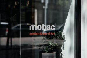 Logo von Mobac mit dem Slogan "motion is our drive" auf einem Schaufenster, umgeben von einer Pflanze in einem Topf.