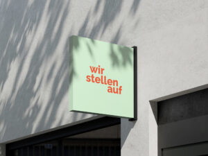 Hängeschild mit der Aufschrift "wir stellen auf" in orange auf grünem Hintergrund, angebracht an einer hellgrauen Wand.