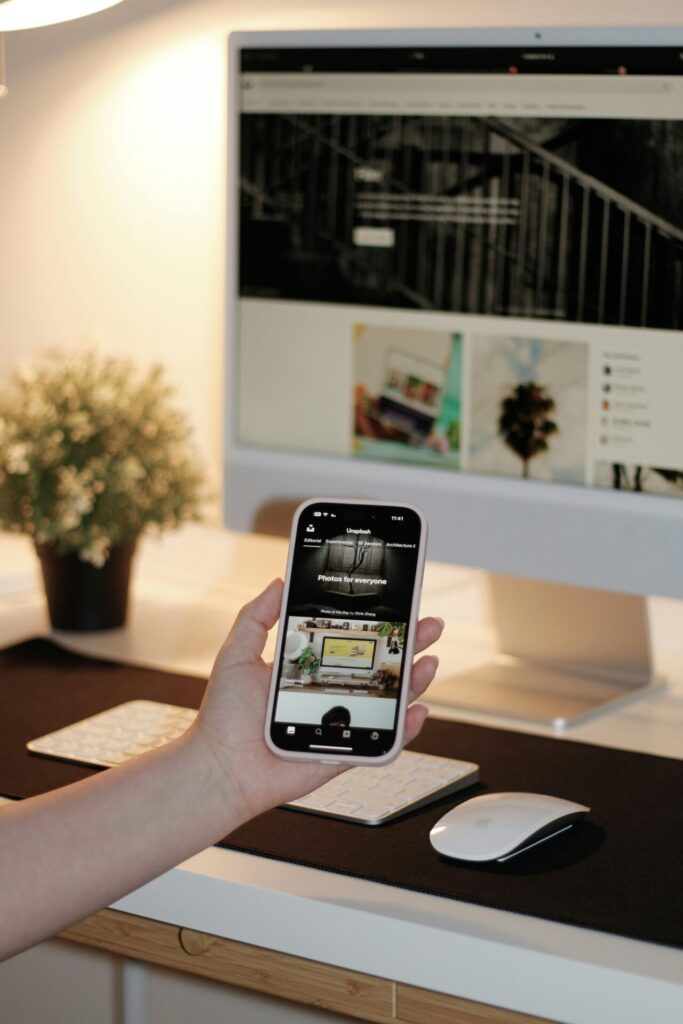 Person hält ein Smartphone in der Hand, auf dem die Unsplash-App geöffnet ist, während im Hintergrund ein Computerbildschirm mit einer Website sichtbar ist. Der Arbeitsplatz ist modern und gut organisiert, mit einer weißen Tastatur und einer Maus auf einem schwarzen Mauspad sowie einer kleinen Pflanze.