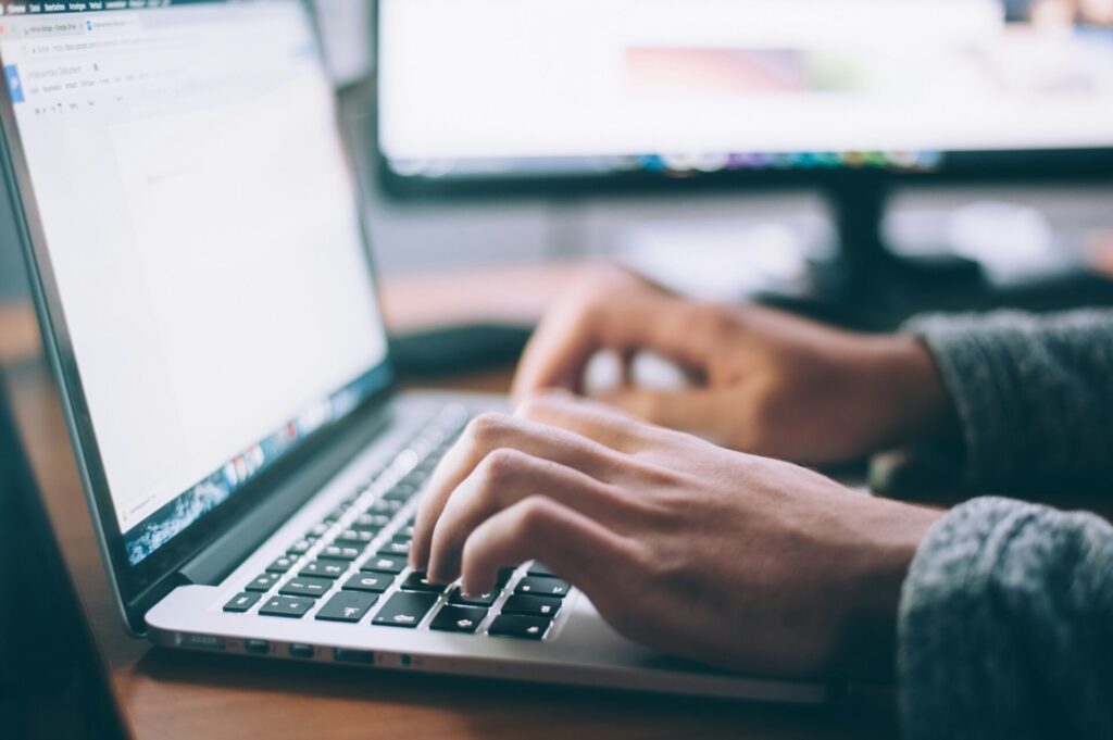 Eine Person tippt auf der Tastatur eines Laptops, während im Hintergrund ein zweiter Monitor sichtbar ist. Der Fokus liegt auf den Händen und der Laptop-Oberfläche, die ein Dokument anzeigen.