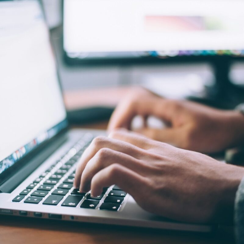 Eine Person tippt auf der Tastatur eines Laptops, während im Hintergrund ein zweiter Monitor sichtbar ist. Der Fokus liegt auf den Händen und der Laptop-Oberfläche, die ein Dokument anzeigen.