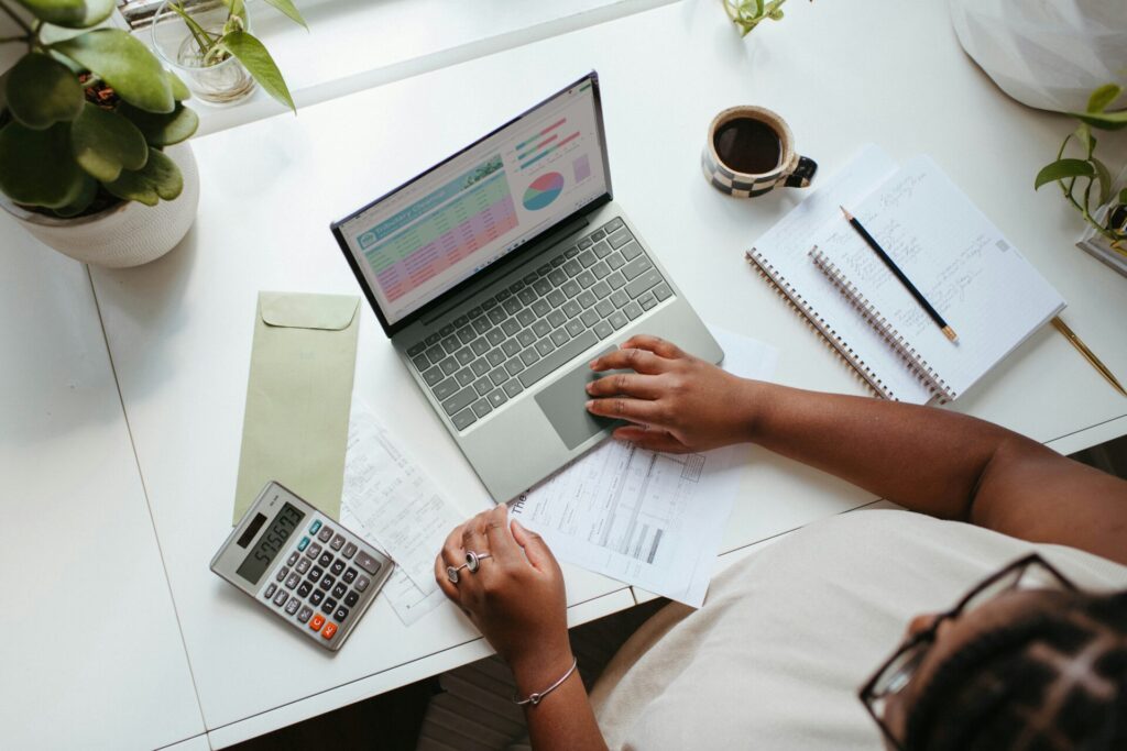 Eine Person arbeitet an einem Laptop, während sie Finanzdaten analysiert. Auf dem Tisch befinden sich eine Schreibmappe, ein Notizbuch mit handschriftlichen Notizen, ein Taschenrechner und eine Tasse Kaffee. Pflanzen dekorieren den Arbeitsplatz und schaffen eine angenehme Atmosphäre für produktives Arbeiten.