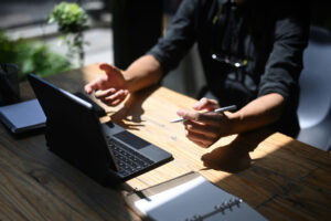 Ein Mensch in schwarzem Hemd sitzt an einem Holztisch und gestikuliert während eines Online-Meetings. Vor ihm steht ein Laptop, daneben liegen ein Notizbuch und ein Stift. Tageslicht strömt durch das Fenster und beleuchtet die Szene.