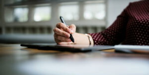 Eine Hand hält einen digitalen Stift und zeichnet auf einem Tablet, während im Hintergrund ein heller Raum mit Fenstern zu sehen ist.