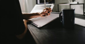 Hand einer Person, die mit einem Stylus auf einem Tablet zeichnet, während ein Laptop und eine Tasse Kaffee auf einem Schreibtisch stehen.