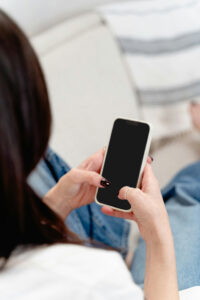 Eine Person sitzt auf einem Sofa und hält ein Smartphone in der Hand, während sie darauf tippt. Die Umgebung ist hell und gemütlich, mit einer Decke im Hintergrund.