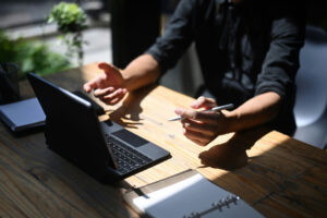 Mann mit Stift in der Hand, der an einem Laptop arbeitet, während er gestikuliert. Auf dem Tisch liegen ein Notizblock und ein Planer. Natürliches Licht strömt durch das Fenster, was eine produktive Arbeitsatmosphäre schafft.