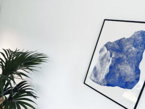 Abstractes Kunstwerk in Blau und Grau, eingerahmt, an einer weißen Wand, mit einer grünen Pflanze im Vordergrund.