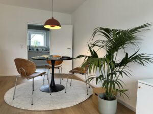 Moderne Essgruppe mit runden Tisch und drei Holzstühlen in einem hellen Raum. Im Hintergrund ist eine Küche sichtbar. Eine große Zimmerpflanze steht in einem grauen Topf neben dem Tisch.
