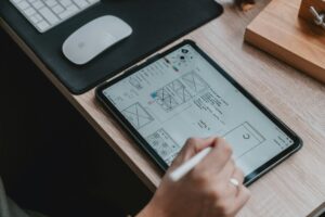 Hand, die mit einem Stylus auf einem Tablet zeichnet, während Wireframes und Notizen für ein Designprojekt angezeigt werden. Im Hintergrund sind eine Computermaus und eine Tastatur sichtbar.
