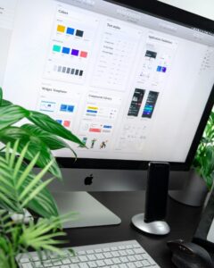 Ein iMac-Bildschirm zeigt Design-Layouts mit Farbpaletten, Textstilen, Widget-Vorlagen und Anwendungshinweisen. Im Vordergrund sind Pflanzen und eine Tastatur sichtbar.