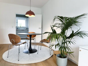 Moderne Essbereich mit einem runden Tisch und drei Holzstühlen, umgeben von einer großen Zimmerpflanze. Im Hintergrund ist eine helle Küche mit Fenster sichtbar. Der Boden ist aus Holz und ein runder Teppich ergänzt die Einrichtung.