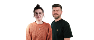 Zwei Personen stehen vor einem schwarzen Hintergrund. Die Frau trägt einen orangefarbenen Pullover und hat eine Brille. Der Mann trägt ein schwarzes T-Shirt und hat einen Bart.