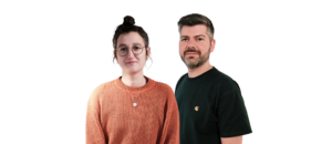 Zwei Personen stehen nebeneinander vor einem neutralen Hintergrund. Die Frau trägt einen orangefarbenen Pullover und hat eine Brille auf, während der Mann ein schwarzes T-Shirt trägt. Im Hintergrund sind Bücherregale zu sehen.