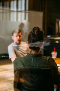 Zwei Personen in einem modernen Büro führen ein Gespräch. Eine Person ist von hinten zu sehen, während die andere im Vordergrund sitzt und aufmerksam zuhört. Auf dem Tisch sind Getränke und Schreibmaterialien zu erkennen.