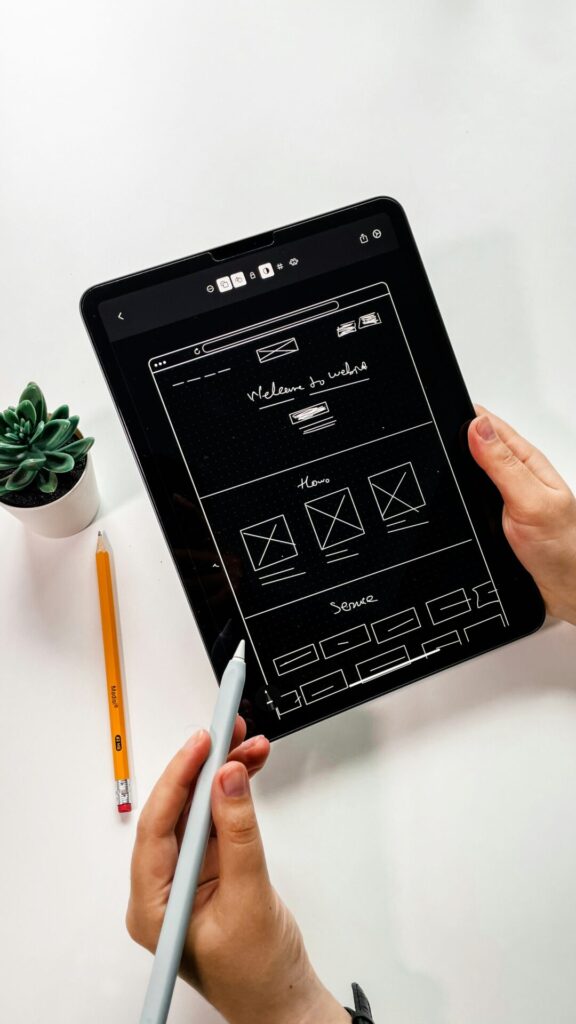 Person hält ein Tablet mit einem Wireframe-Design für eine Website, darauf sind verschiedene Layouts und Navigationselemente skizziert. Neben dem Tablet liegen ein Bleistift und ein stylischer Stift. Im Hintergrund ist eine kleine Pflanze zu sehen.