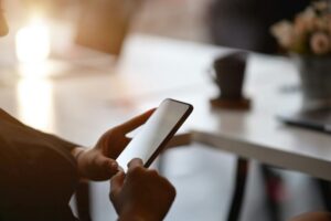 Person hält ein Smartphone in der Hand und tippt darauf, während im Hintergrund ein Tisch mit einer Tasse Kaffee und einer Blume zu sehen ist. Das Bild vermittelt eine entspannte Arbeitsatmosphäre.