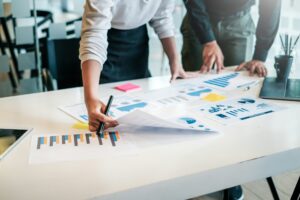 Zwei Personen analysieren gemeinsam Diagramme und Grafiken auf einem Tisch, während eine Person mit einem Stift Notizen macht. Auf dem Tisch liegen verschiedene Dokumente mit Datenvisualisierungen, Post-it-Zettel und ein Laptop.