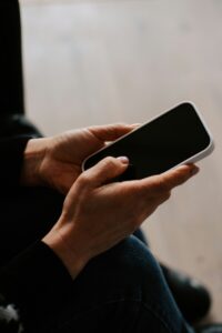 Person hält ein Smartphone in der Hand, während sie auf einem Stuhl sitzt. Das Handy hat einen weißen Rahmen und einen schwarzen Bildschirm. Die Umgebung ist hell und neutral gestaltet.