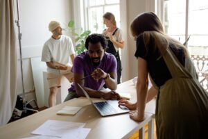 Eine Gruppe von kreativen Fachleuten arbeitet zusammen an einem Projekt in einem hellen, modernen Büro. Ein Mann erklärt eine Idee am Laptop, während eine Frau ihm aufmerksam zuhört. Im Hintergrund stehen weitere Teammitglieder, die Notizen machen.