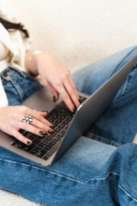 Eine Person mit dunklem Nagellack tippt auf einem Laptop, während sie in bequemen Jeans sitzt. Die Hände sind mit Ringen geschmückt und der Hintergrund ist hell und neutral.