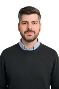 Porträt eines Mannes mit dunklem Pullover und hellblauem Hemd, der direkt in die Kamera blickt. Der Hintergrund ist weiß, was den Fokus auf sein Gesicht und seine gepflegte Erscheinung legt.