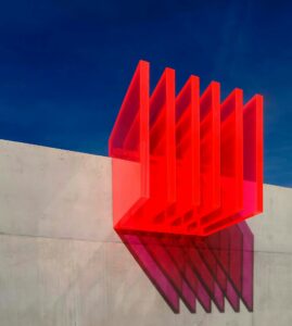 Rote transparente Strukturen, die an einer grauen Wand hängen, werfen Schatten auf die Oberfläche. Der klare blaue Himmel im Hintergrund verstärkt die visuelle Wirkung der modernen Architektur.