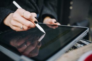 Eine Hand mit schwarzem Nagellack hält einen Stift und tippt auf einem Tablet-Bildschirm, während der Text auf dem Display reflektiert wird.