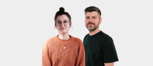 Portrait eines Paares, das in lässiger Kleidung vor einem einfarbigen Hintergrund steht. Die Frau trägt einen orangefarbenen Pullover und hat eine Brille auf, während der Mann ein schwarzes T-Shirt trägt.