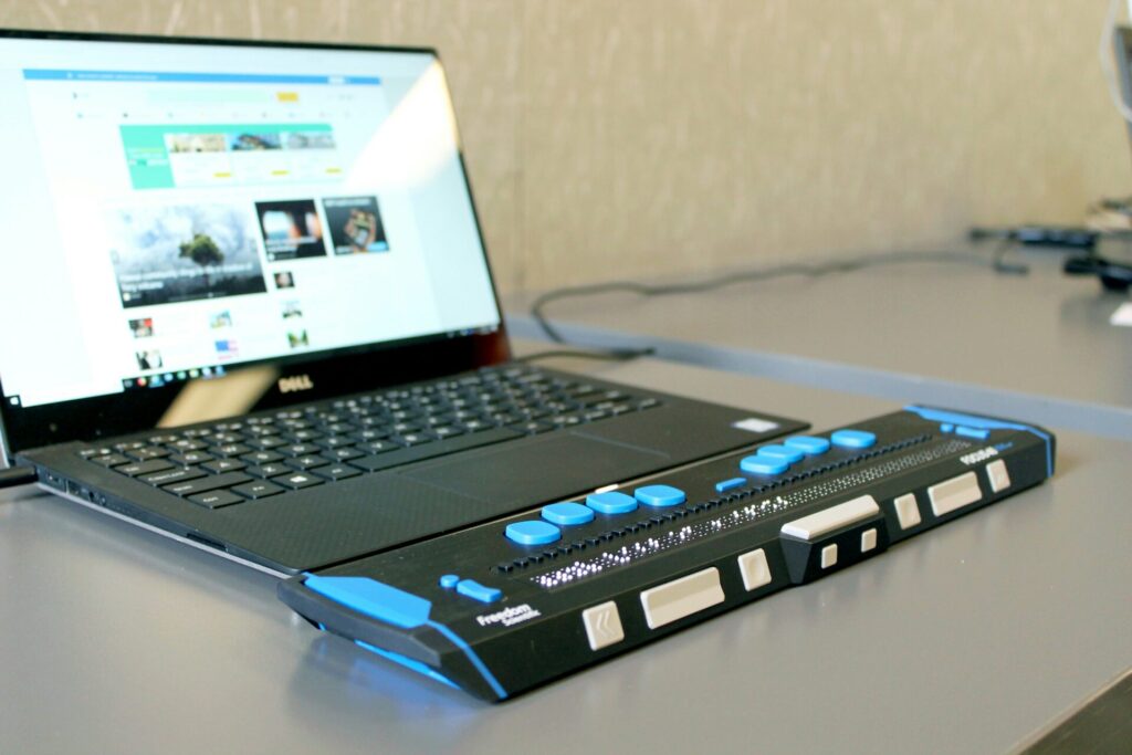 Ein Laptop von Dell steht auf einem Tisch, neben dem eine Braillezeile mit blauen Tasten und einer Punktmatrix platziert ist. Der Laptop zeigt eine Webseite an, die Informationen und Medieninhalte präsentiert.