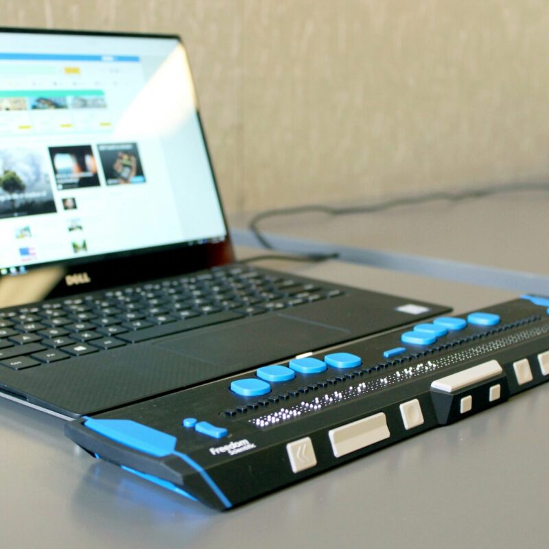 Ein Laptop von Dell steht auf einem Tisch, neben dem eine Braillezeile mit blauen Tasten und einer Punktmatrix platziert ist. Der Laptop zeigt eine Webseite an, die Informationen und Medieninhalte präsentiert.