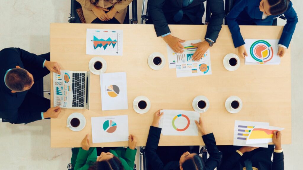 Teammeeting mit Diagrammen und Statistiken auf einem Tisch, umgeben von Tassen Kaffee. Mitarbeiter analysieren Daten auf Laptops und Papierunterlagen.
