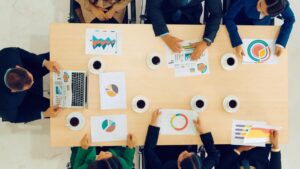 Teammeeting mit Diagrammen und Statistiken auf einem Tisch, umgeben von Tassen Kaffee. Mitarbeiter analysieren Daten auf Laptops und Papierunterlagen.